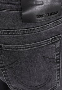 Jean en denim noir avec une étiquette de taille en cuir embossée avec "True Religion". Détails de couture subtils et design de poche arrière visibles.