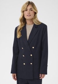 Blazer bleu marine croisé avec boutons dorés, grandes poches avant et coupe ajustée. Le tissu semble lisse avec un design structuré.