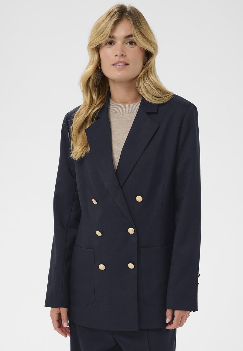 Blazer bleu marine croisé avec boutons dorés, grandes poches avant et coupe ajustée. Le tissu semble lisse avec un design structuré.