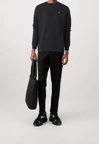 Sudadera negra con un logo amarillo, pantalones negros y bolso tote acolchado negro. Zapatillas deportivas negras con detalles en blanco. Diseño sencillo.