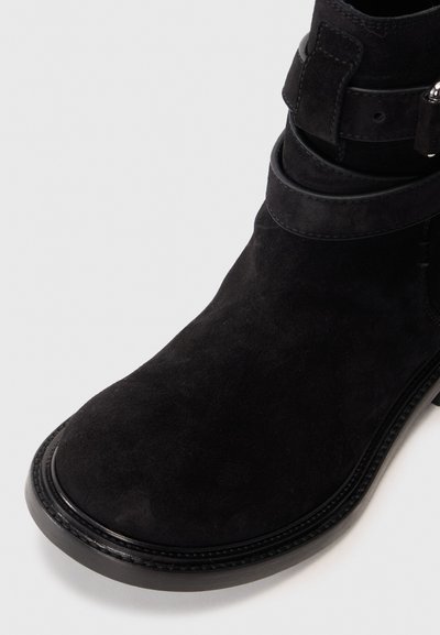 Botă ankle din suede negru, cu o textură netedă, vârf rotund și o curea decorativă în jurul gleznei. Are o talpă din cauciuc rezistentă.
