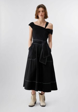 Ženska v črni obleki brez ramen z zavezanim pasom in žepi, obuta v svetle gležnjarje, stoji pred enobarvnim ozadjem.