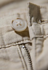 Lichtbeige stof met een cirkelvormige, roomkleurige knop met vier gaten en een zilveren rits nabij de rand met zichtbare stiksels.