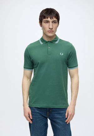 Mężczyzna w zielonej koszulce polo z krótkim rękawem, z białym paskowanym kołnierzykiem i krawędziami rękawów, w połączeniu z niebieskimi dżinsami, stoi na jednolitym tle.