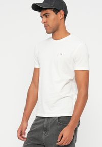 Λευκό βαμβακερό t-shirt με κοντά μανίκια, στρογγυλή λαιμόκοψη και μικρή ετικέτα λογότυπου στο στήθος. Το μοντέλο φοράει σκούρο καπέλο και γκρι παντελόνι.