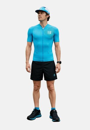 Homme portant un maillot de cyclisme sportif turquoise, un short noir, des chaussures de course bleu et noir, une casquette bleue et une montre noire, debout et regardant vers la droite.
