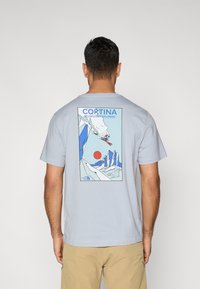 Ανοιχτό μπλε βαμβακερό μπλουζάκι με σχέδιο στην πλάτη που απεικονίζει βουνά, έναν σκιέρ και έναν κόκκινο ήλιο. Το κείμενο αναγράφει «CORTINA NEVER STOP EXPLORING».