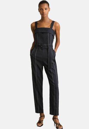 Zwarte jumpsuit met brede bandjes, vierkante halslijn, ceintuur op de taille en contrasterende witte stiksels. Afgelotte pijpen en zijzakken.