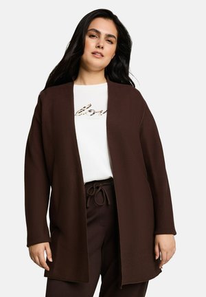 Kobieta nosząca brązowy sweter rozpinany na przodzie, na biały T-shirt z napisem oraz dopasowane brązowe spodnie ze ściągaczem w pasie.