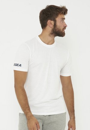 Man met baard die een wit t-shirt met korte mouwen draagt met zwarte tekst "LEGEA" op de linkermouw en grijze broek, kijkt naar rechts.