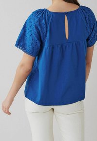 Blusa di cotone blu con maniche ad occhiello ricamate, scollo tondo, apertura a chiave sul retro e vestibilità ampia, abbinata a pantaloni chiari.