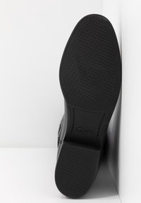 Chaussure en cuir noir avec une semelle en caoutchouc texturée, dotée d'un bout arrondi et de détails de couture subtils. Nom de la marque embossé sur la semelle.