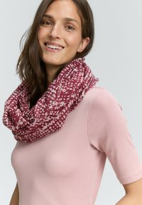 Frau, die lächelt, trägt ein rosa Kurzarmoberteil und einen gemusterten burgunderfarbenen und weißen Infinity-Schal vor einem einfarbigen hellen Hintergrund.