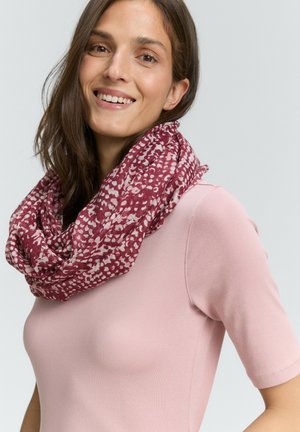 Frau, die lächelt, trägt ein rosa Kurzarmoberteil und einen gemusterten burgunderfarbenen und weißen Infinity-Schal vor einem einfarbigen hellen Hintergrund.