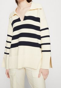 Cremfarbener Pullover mit marineblauen horizontalen Streifen, tiefem V-Ausschnitt, geripptem Saum und seitlichen Schlitzen. Hergestellt aus strukturiertem Material.