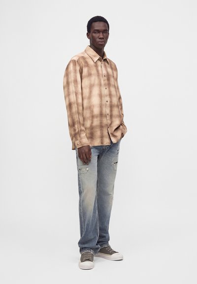 Jeune homme debout portant une chemise beige à carreaux boutonnée, un jean bleu délavé déchiré et des baskets grises sur un fond blanc uni.