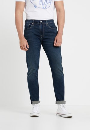 Mörkblå slim-fit jeans med en slät textur, fem fickor och uppvikta nederdelar, tillsammans med en vit grafisk t-shirt och vita sneakers.
