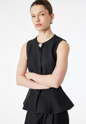 Femme aux cheveux attachés portant un haut noir sans manches avec un bouton argenté, les bras croisés, sur un fond blanc uni.