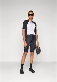 Μαύρη φανέλα ποδηλασίας πάνω από λευκό βασικό στρώμα, μαύρο σορτς, μαύρες κάλτσες και παπούτσια· φορά γυαλιά ηλίου και κρατά κράνος.