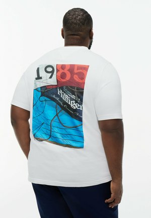 Homme portant un t-shirt blanc avec un grand graphique d'un voilier rouge, blanc et bleu et le texte "1985 Tommy Hilfiger" dans le dos.