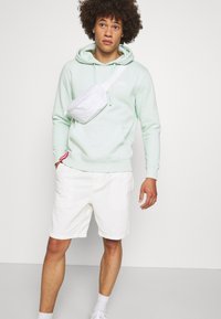 Felicidade é um hoodie verde menta claro com cordões, bolso canguru, e uma bolsa crossbody branca usada no peito. Calções brancos e ténis.