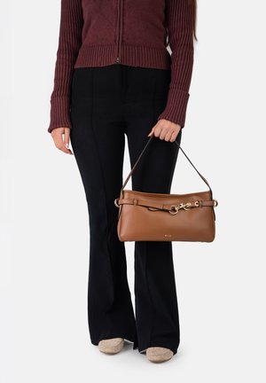 Žena u rebrastom smeđem džemperu i crnim hlačama drži smeđu kožnu torbicu sa zlatnim okovima i kratkom naramenicom.