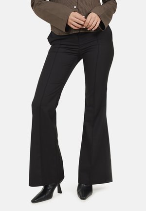 Vrouw draagt zwarte flared broek, zwarte schoenen met hak en een bruin gestreepte lange mouw top met knoopmanchetten.