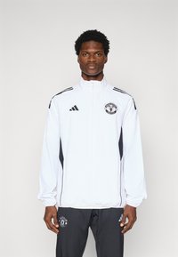 White zip-up jacket with black side panels, featuring the Manchester United logo on the chest and three black stripes on each shoulder.