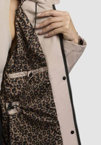 Veste extérieure beige clair avec une doublure à imprimé léopard. Les caractéristiques incluent une fermeture éclair, des boutons-pression et une poche intérieure avec un bouton.