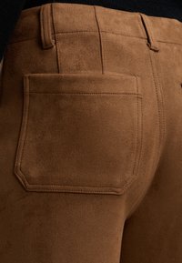 Καφέ παντελόνια από σουέτ που δείχνουν το ράψιμο στην πίσω τσέπη και τις θηλιές της ζώνης, με ένα μαύρο τοπ που φαίνεται μερικώς πάνω από τη μέση.