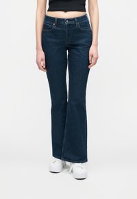 Άτομο που φοράει σκούρα μπλε τζιν bootcut, μαύρο κοντό τοπ και λευκά platform αθλητικά παπούτσια, που στέκεται μπροστά σε λευκό απλό φόντο.