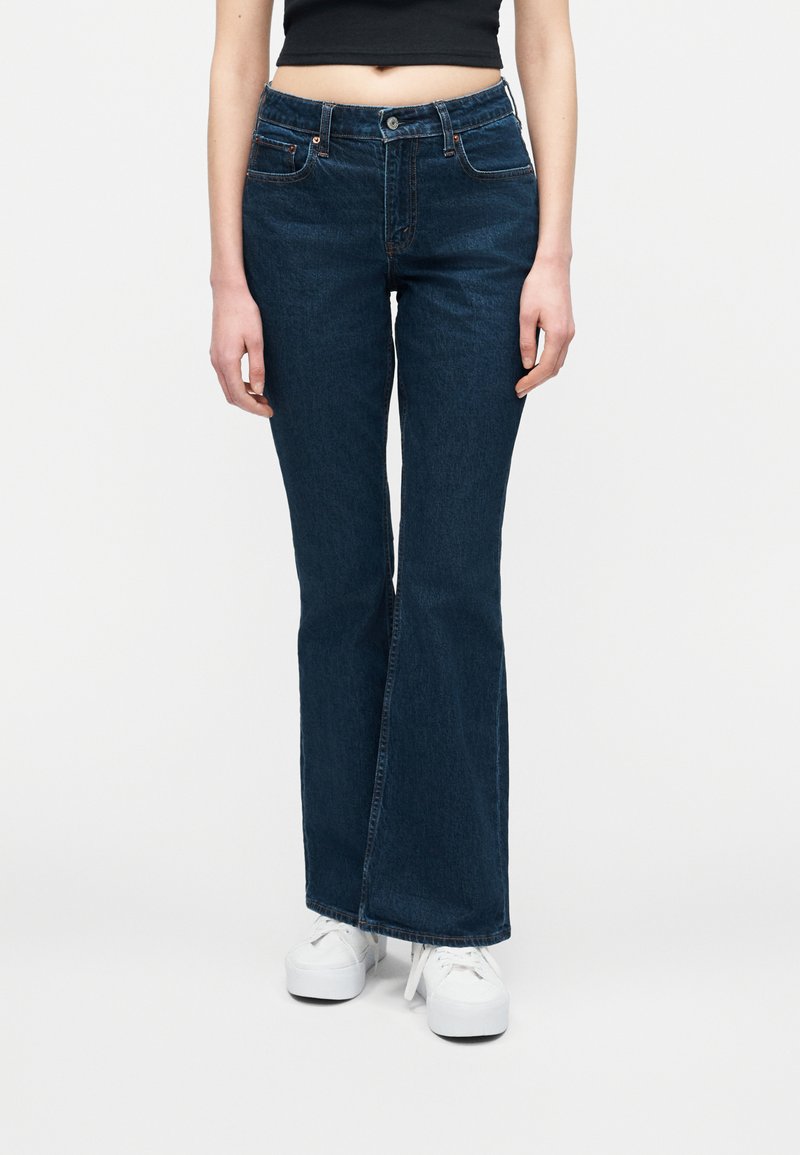 Άτομο που φοράει σκούρα μπλε τζιν bootcut, μαύρο κοντό τοπ και λευκά platform αθλητικά παπούτσια, που στέκεται μπροστά σε λευκό απλό φόντο.