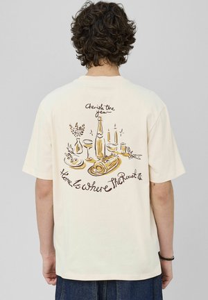 T-shirt couleur crème avec un graphisme au dos représentant de la nourriture, des boissons, et le texte "Chérissez l'année, la maison est là où se trouve le rôti."