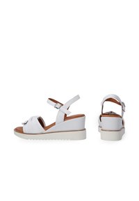 Witte wedge-sandalen met een knoopontwerp aan de voorkant, verstelbaar enkelbandje en een gestructureerde witte zool. Bruine binnenzool voor contrast.