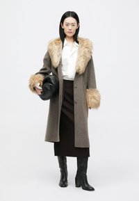 Lang grå frakke med faux fur krave og manchetter, knaplukning foran. Modellen holder en sort lædertaske. Nederdel og knæ-høje sorte støvler fuldender looket.