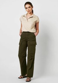 Beige Hemd mit Kragen, kombiniert mit olivgrünen Cargo-Hosen mit großen Taschen; glattes Material; lockere Passform; schwarze Riemchensandalen.