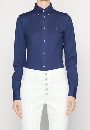 Persoon draagt een marineblauw overhemd met kraag en witte hoge taillebroeken met meerdere zichtbare knopen aan de voorkant.