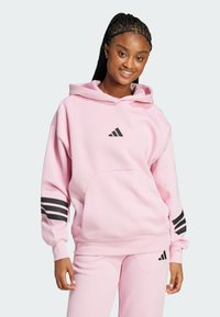 Ροζ φούτερ με κουκούλα, με μαύρο λογότυπο Adidas και τρεις ρίγες στα μανίκια. Φτιαγμένο από μαλακό, υφασμάτινο υλικό. Διαθέτει μπροστινή τσέπη.