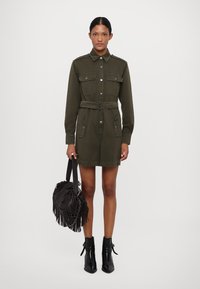 Vestito camicia verde oliva con tasche frontali, chiusura a bottoni e una cintura in vita, abbinato a una borsa nera con frange e stivaletti alla caviglia.