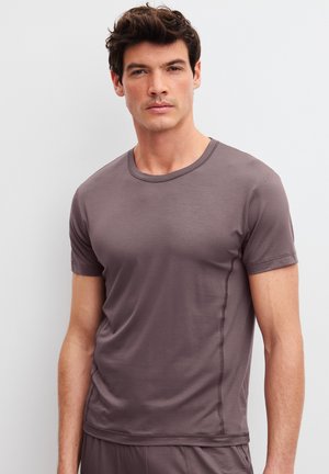 Man die een aansluitend donkergrijs T-shirt met korte mouwen draagt, staat tegen een effen witte achtergrond.