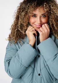 Lichtblauwe gewatteerde jas met een quilted patroon, hoge kraag en zwarte drukknoopjes, snugly om de nek gedragen. Zachte, gladde textuur.