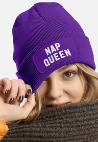 Purple knit beanie with a wide patch labeled "NAP QUEEN" in bold white letters. The fabric has a ribbed texture, and it's snug-fitting.