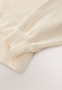 Manches de sweat-shirt beiges avec poignets côtelés, coutures piquées et texture douce du tissu, présentées sur une surface claire.