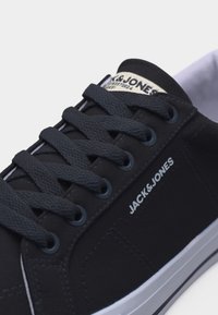 Μαύρο sneaker χαμηλού προφίλ με σκουρόχρωμα κορδόνια και λευκή επιγραφή "JACK & JONES" στο πλάι και στη γλώσσα, σε λευκό φόντο.