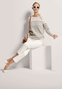 Gestreifter Strickpullover in Beige und Grau, cremefarbene Hose, weiße spitze Ballerinas und übergroße weiße Sonnenbrille, sitzend auf einer geometrischen Plattform.