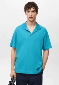 Turkizna polo majica s kratkimi rokavi in ovratnikom, teksturirana blaga, sproščen kroj in podrobnost stranske žepe. Model drži torbo v eni roki.