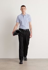 Hellblaues Poloshirt mit weißen Akzenten, schwarze plissierte Hose und schwarze Schuhe, während das Modell eine schwarze Tasche hält. Einfaches, entspanntes Design.