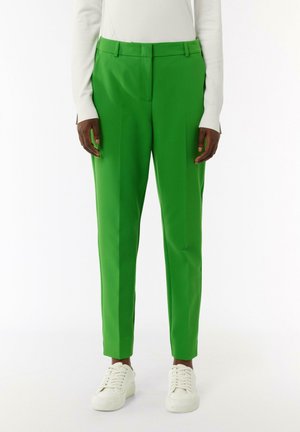 Grüne maßgeschneiderte Hose aus glattem Stoff, mit geradem Schnitt und zwei vorderen Falten, kombiniert mit weißen Sneakers.