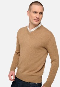 Beige V-Ausschnitt-Pullover aus weichem Strickmaterial, mit gerippten Bündchen und geradem Saum, getragen über einem weißen Hemd mit Kragen.