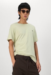 Light green cotton t-shirt with a crew neck, short sleeves, and a small embroidered logo on the chest, paired with dark brown shorts.